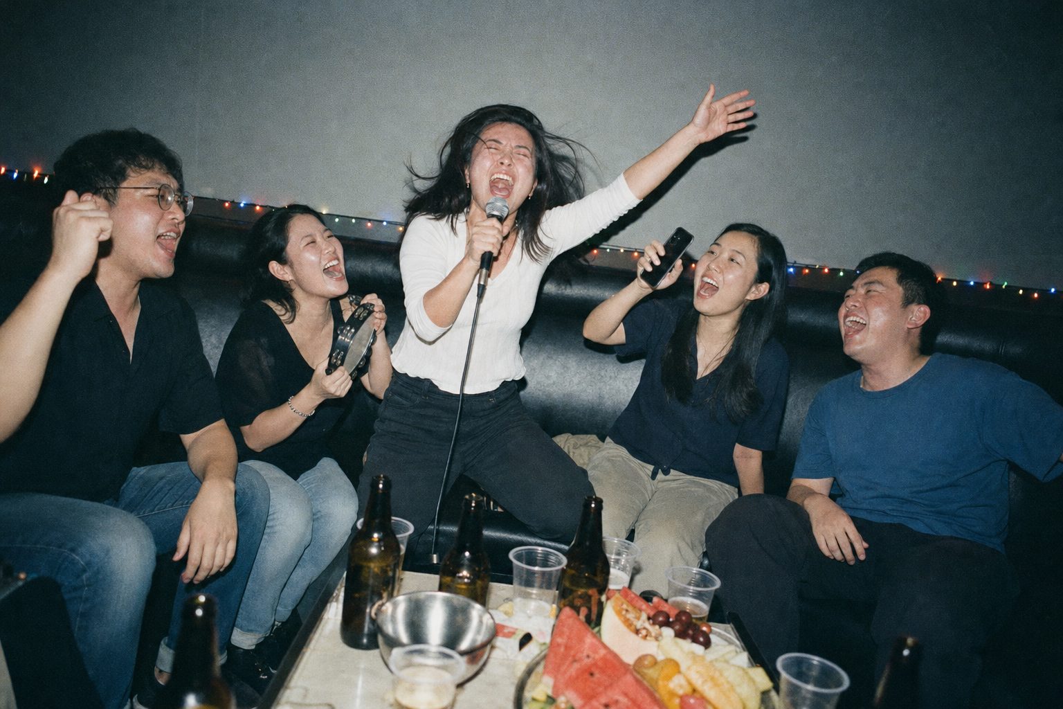 A woman kneeling up on a karaoke sofa with the microphone, eyes shut, hair flying, one arm thrown up, while four friends either side of her sing the chorus back